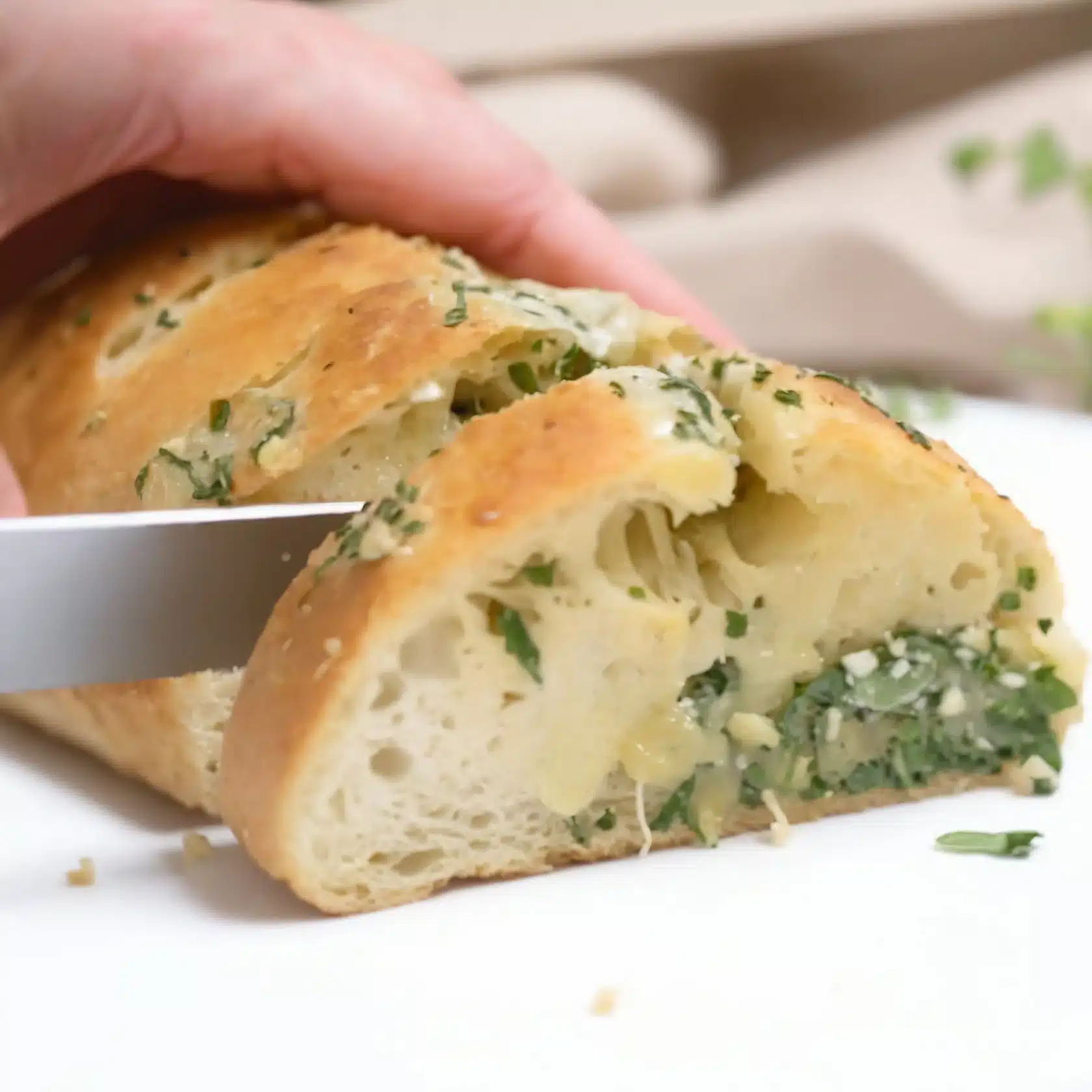 A hand using a knife to slice through a loaf of garlic bread revealing a cheesy, herb-filled interior.